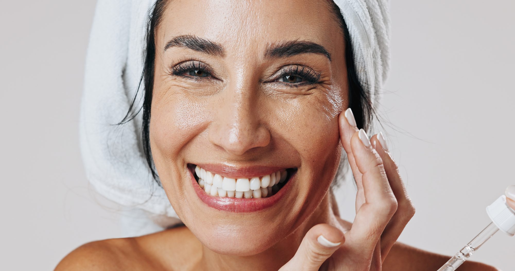 Smiling woman applying skincare serum to her face as part of an anti-aging routine.