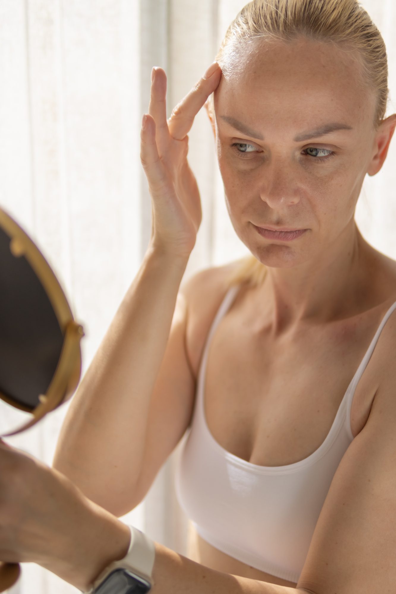 Woman looking in a mirror while touching her forehead and examining her skin.